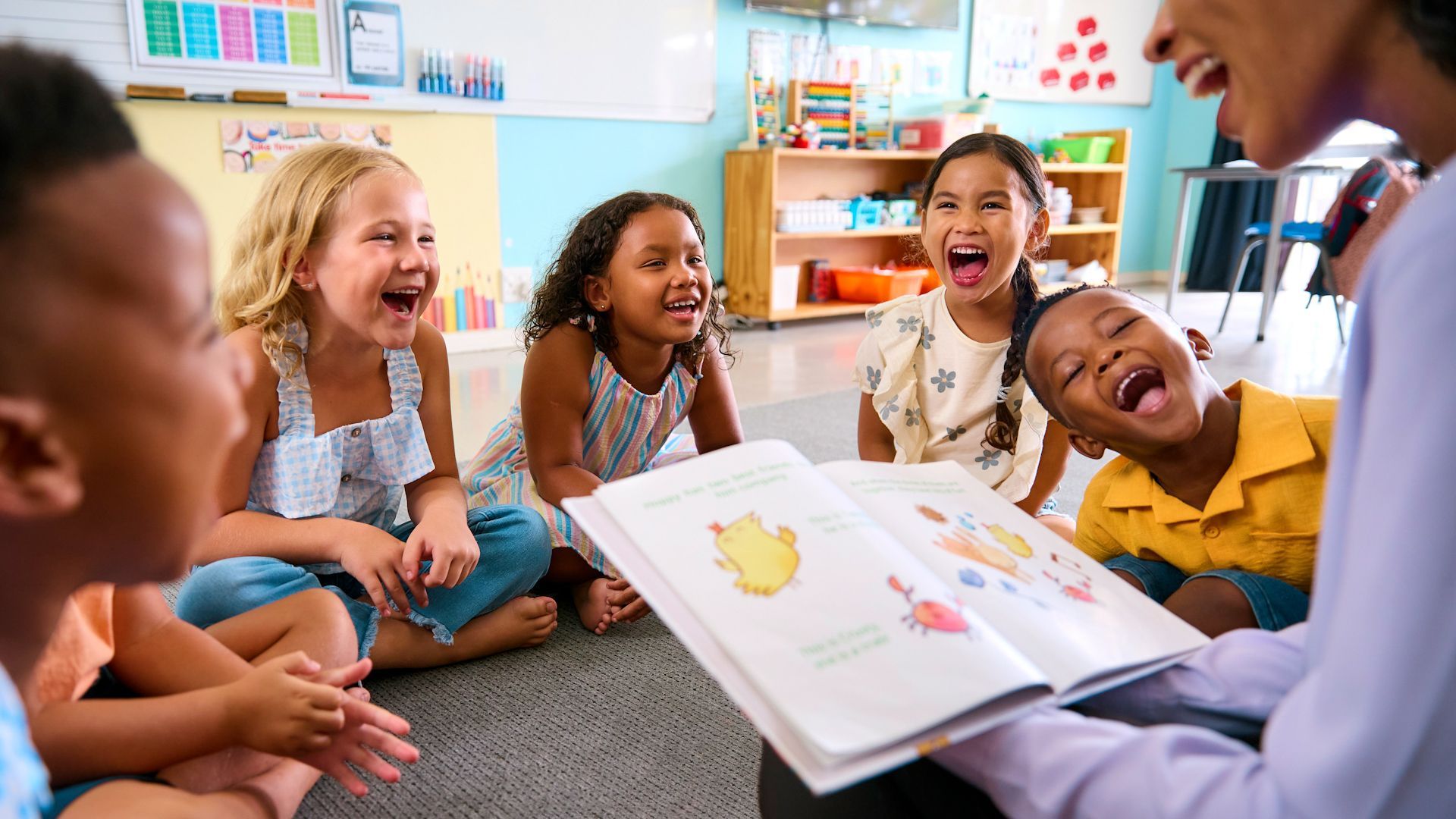 Children at a table in a brightly colored classroom are eating fruit. One child gives an “okay” hand gesture.