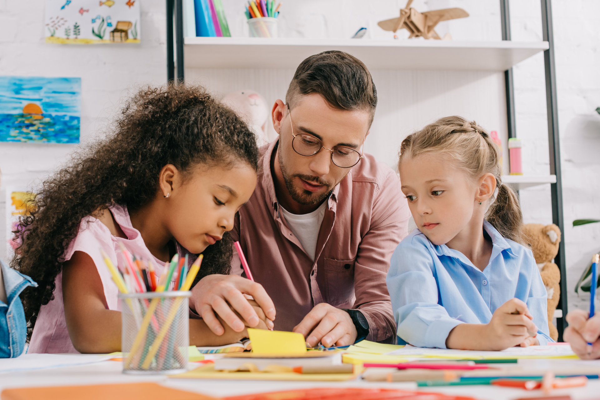 A male teacher with glasses assists two young girls at a table with art supplies. They're focused on a yellow paper.