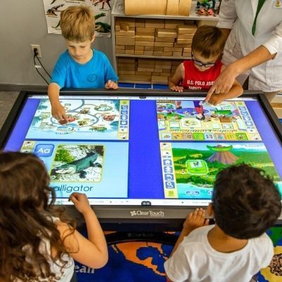 Children at a large interactive table in a classroom are learning with a teacher. They are looking at different educational games.