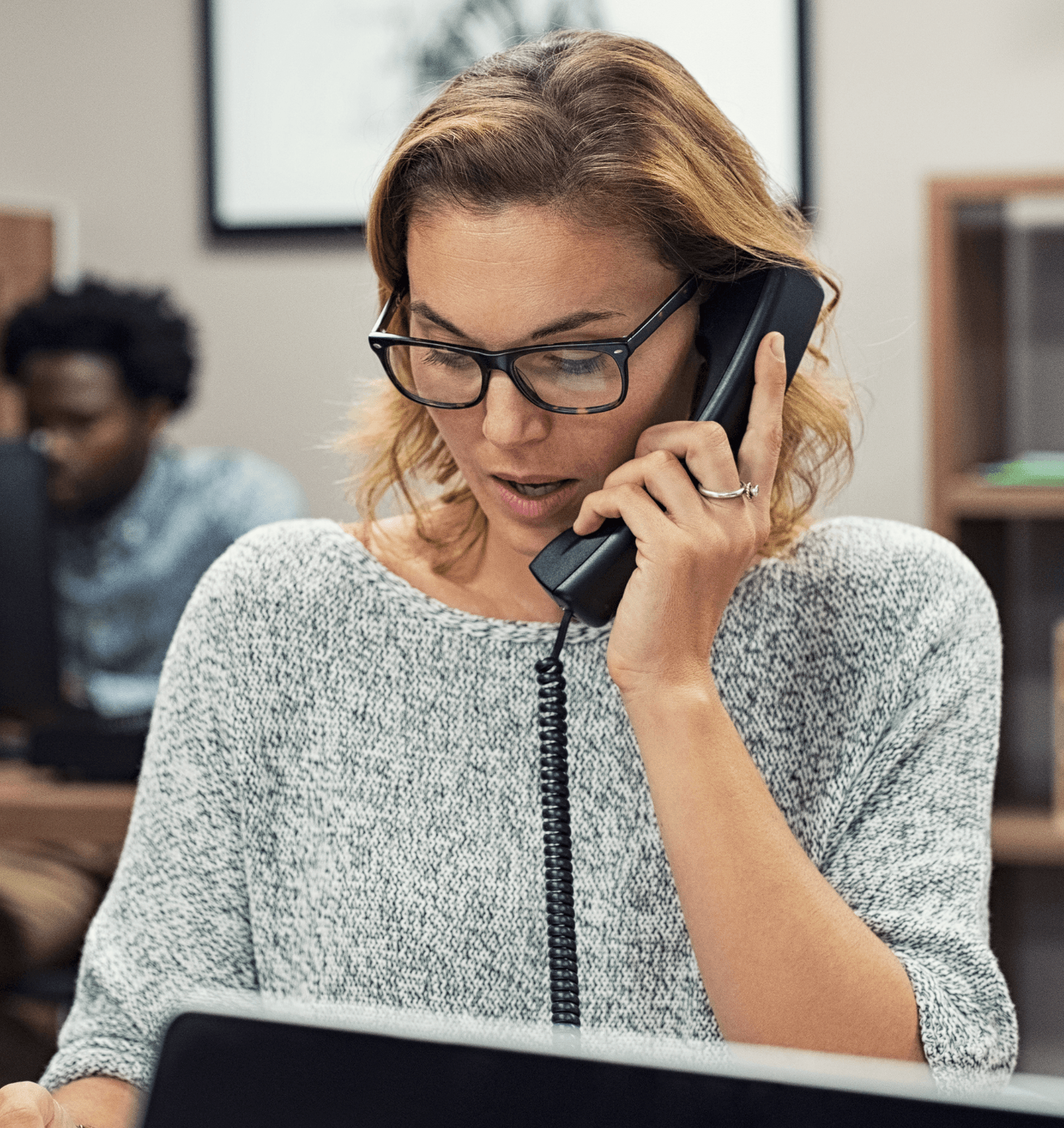 Woman with glasses talks on a landline phone in an office. She wears a gray sweater, looking focused. A person is seated in the background.