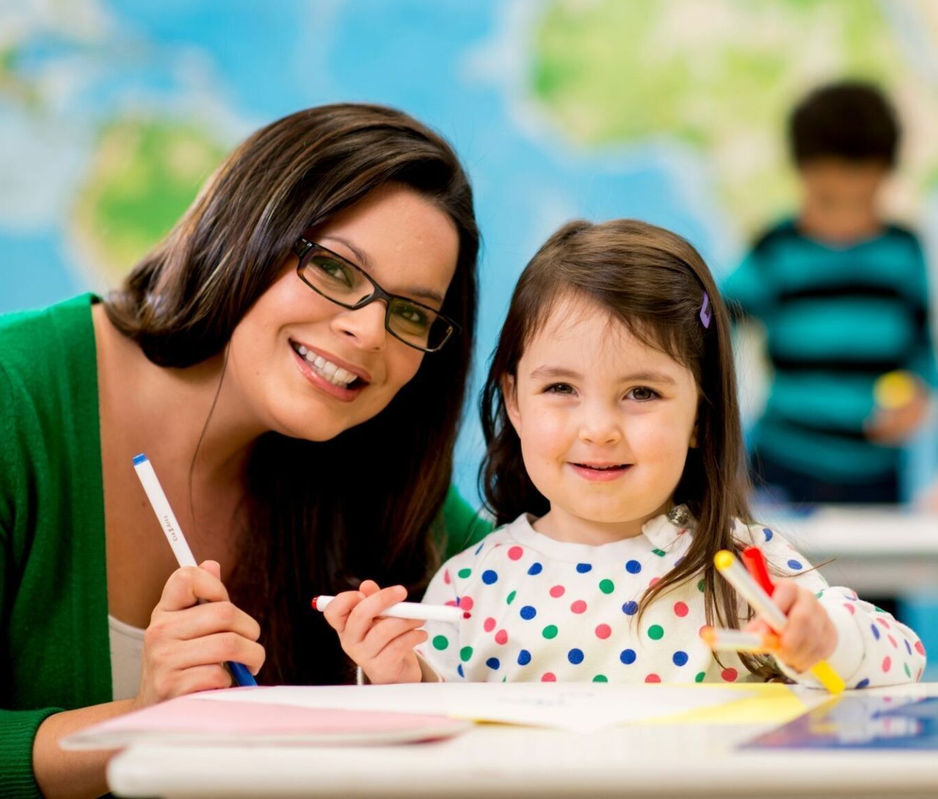 Teacher and young girl smiling while drawing with markers in a classroom; a child is visible in the background.