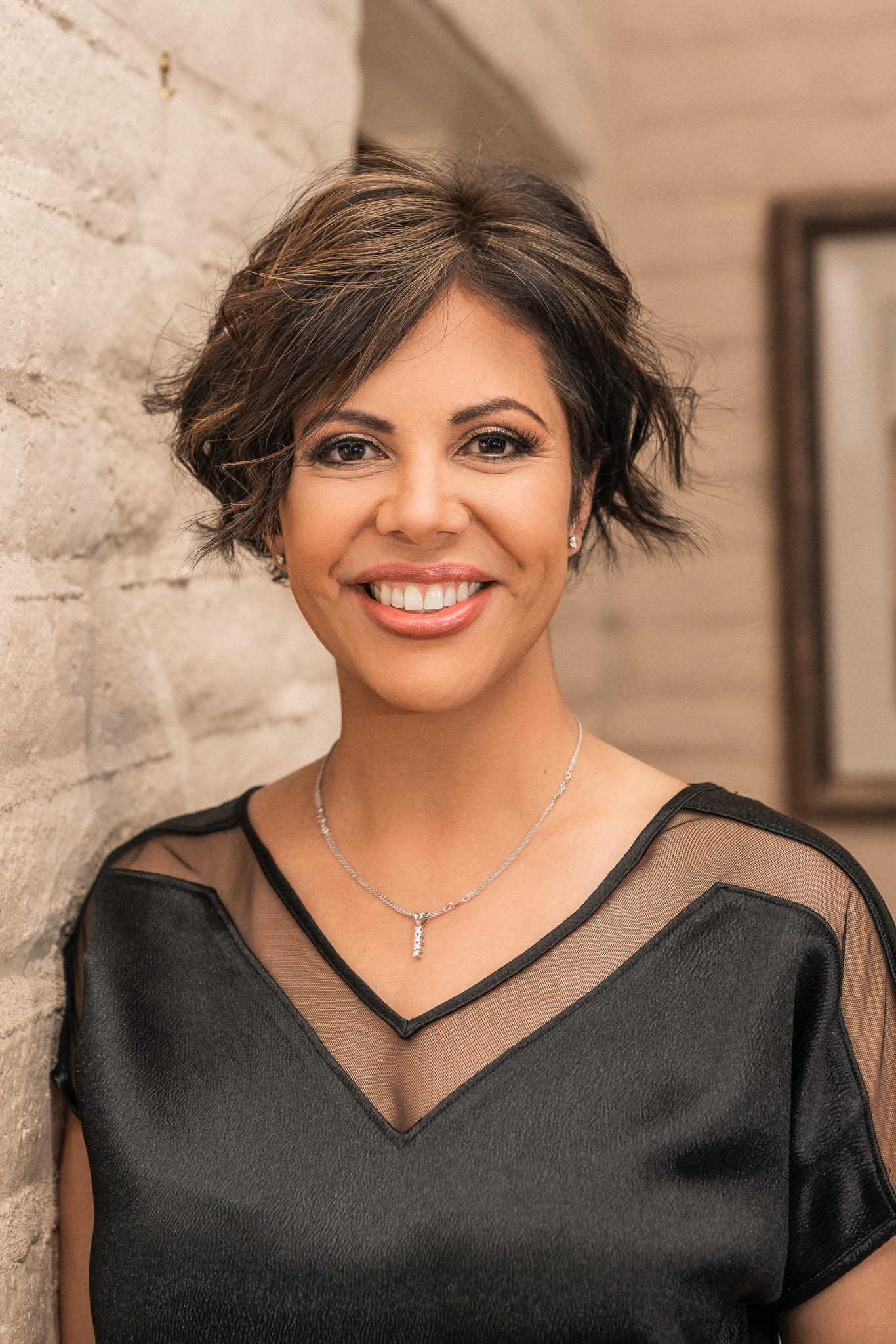 A woman in a black dress is smiling and leaning against a wall.