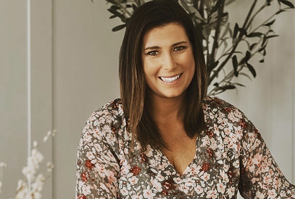 A woman in a black dress is smiling and leaning against a wall.