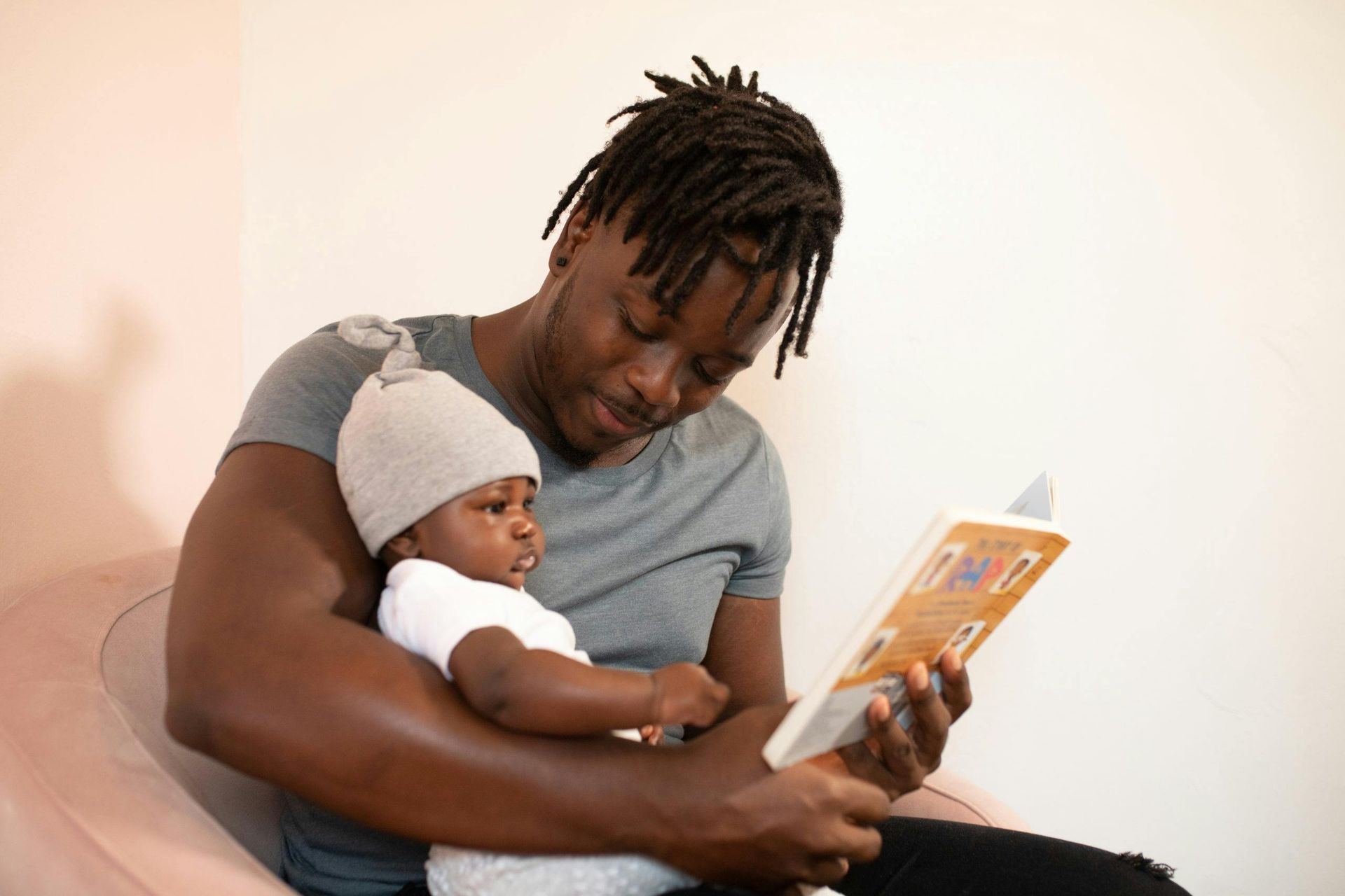 A Black man reads a book to a baby in his arms. The baby wears a hat and looks at the book. They're in a room with a pink couch.