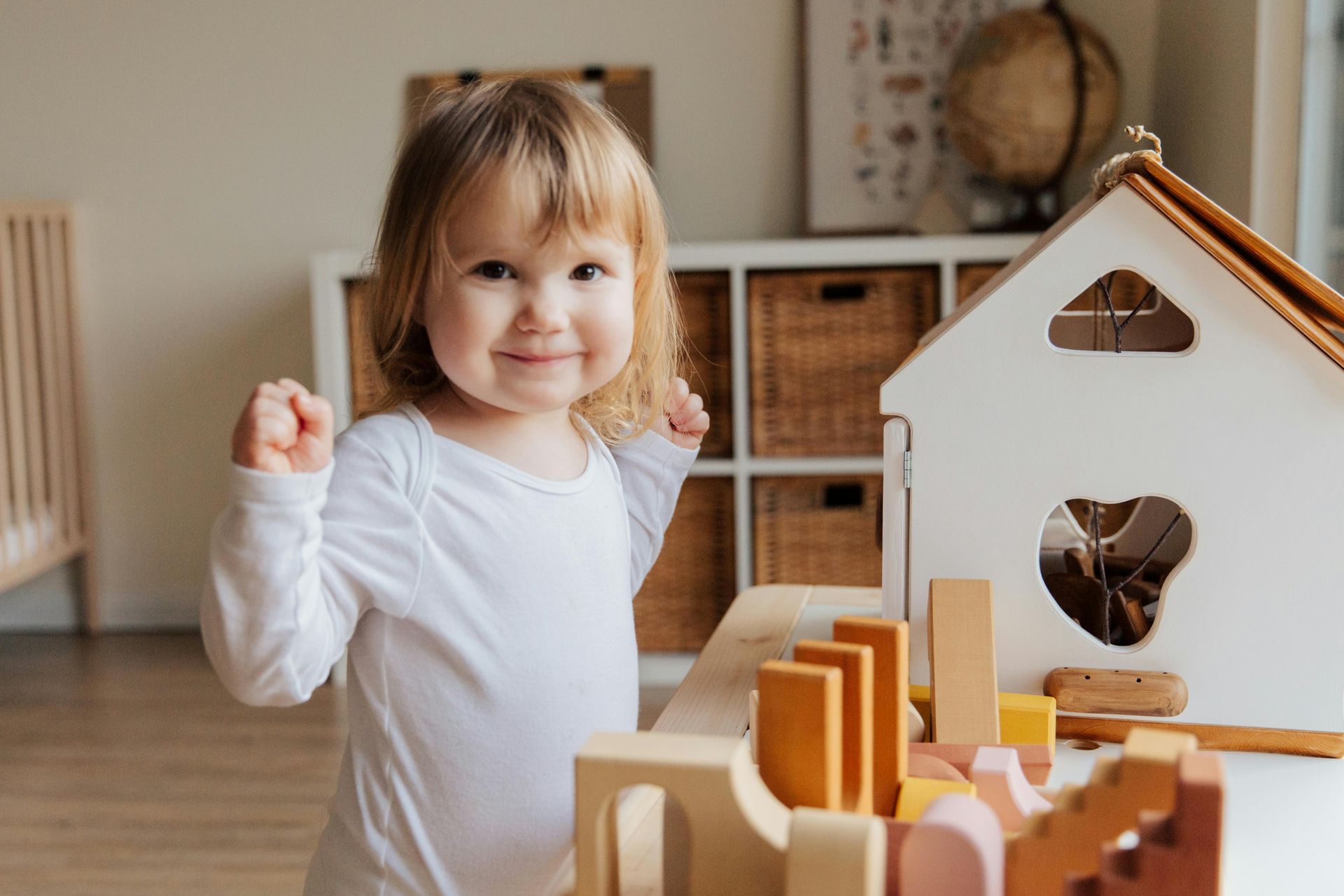 A smiling toddler in a white shirt raises fists near a wooden toy house and blocks in a playroom.