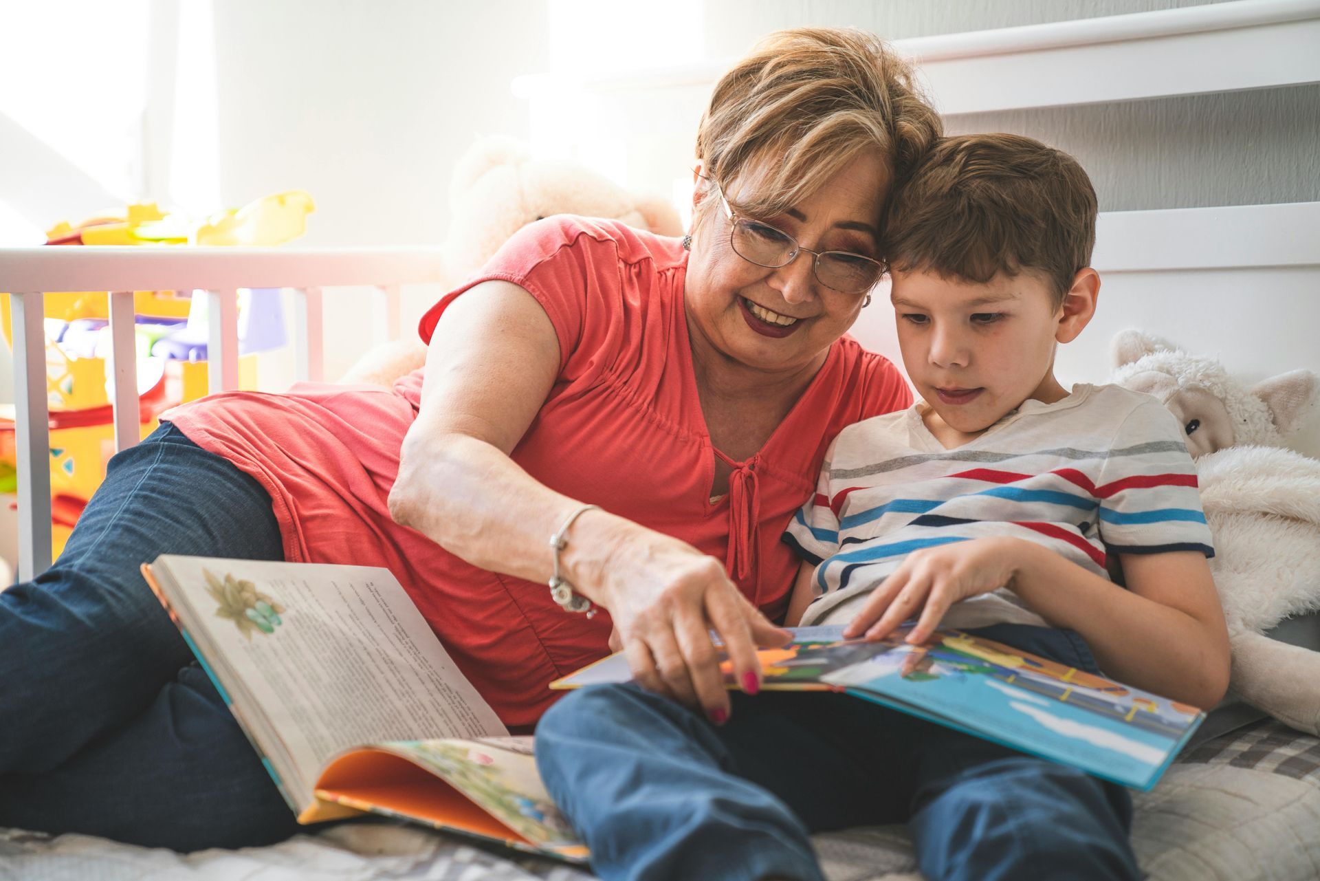 Grandmother and young boy reading a book together on a bed. They are smiling and pointing at the book.