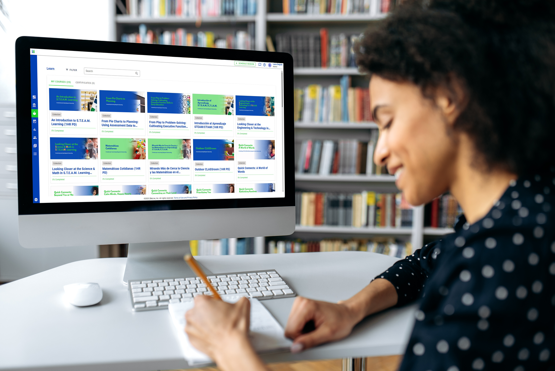 Woman at desk, looking at computer screen, writing in notebook; bookshelves in background.