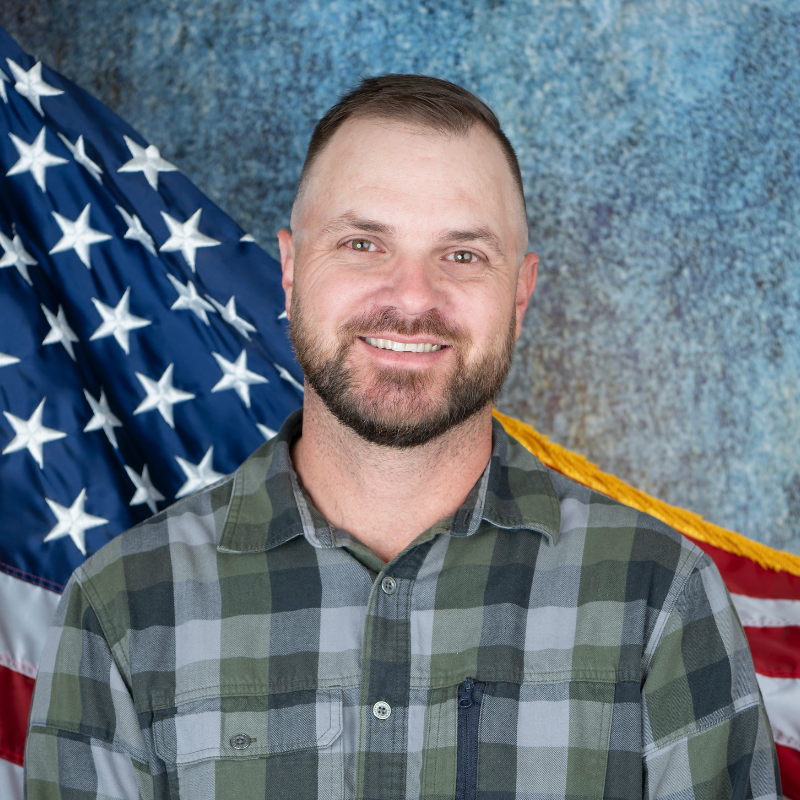 A man with a short beard smiles, wearing a green and gray plaid shirt in front of an American flag.