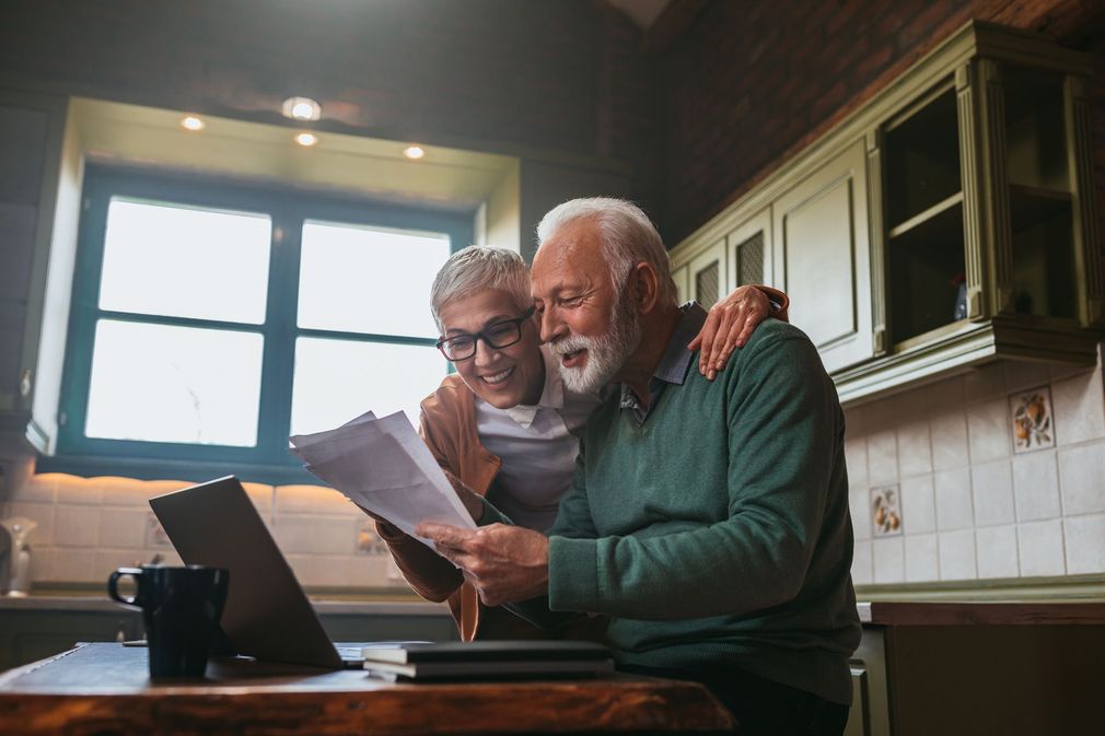 A couple of older men are sitting at a table looking at papers and a laptop.