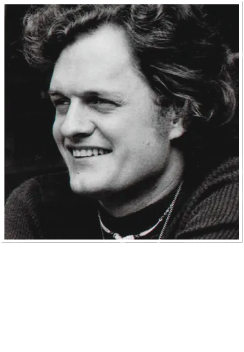 Black and white close-up portrait of a person with wavy hair smiling, wearing a dark sweater and a thin chain necklace.