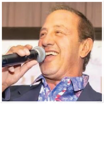 A person wearing a blue patterned shirt and dark jacket smiles while speaking into a handheld microphone.