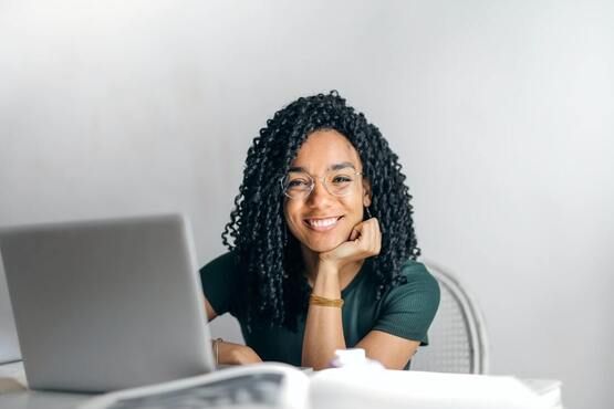 Woman with glasses smiles, resting chin on hand, looking at camera by a laptop.