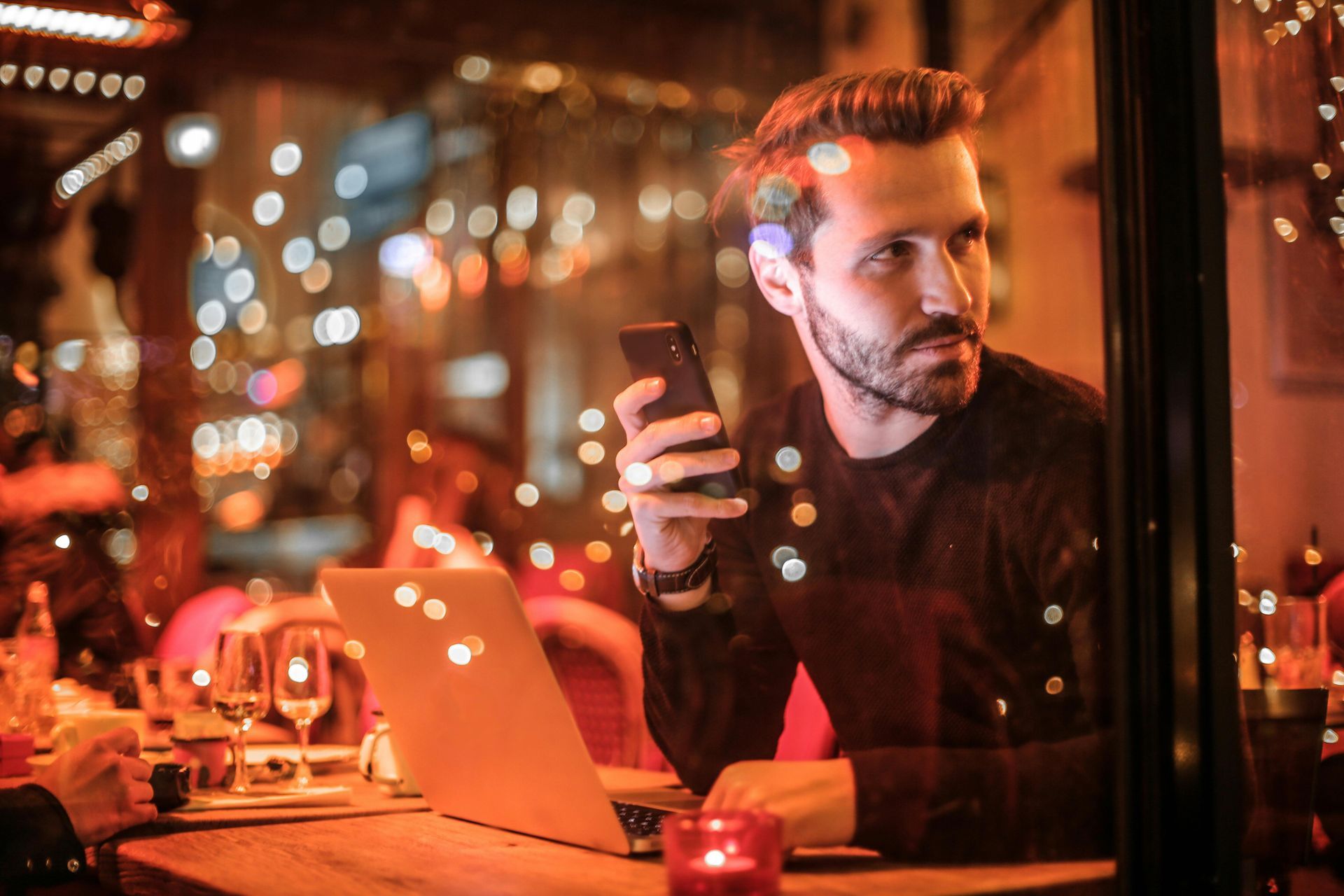 Man at table with laptop, holding phone, looking out window at city lights.