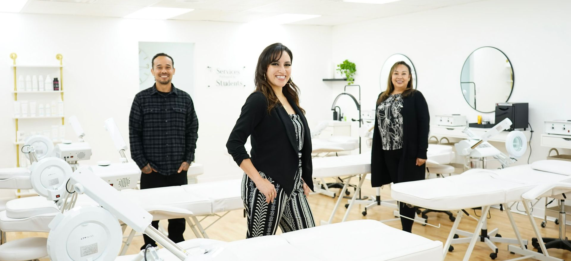 Three licensed estheticians  stand in a bright, modern esthetics class room; white treatment beds and equipment fill the room.