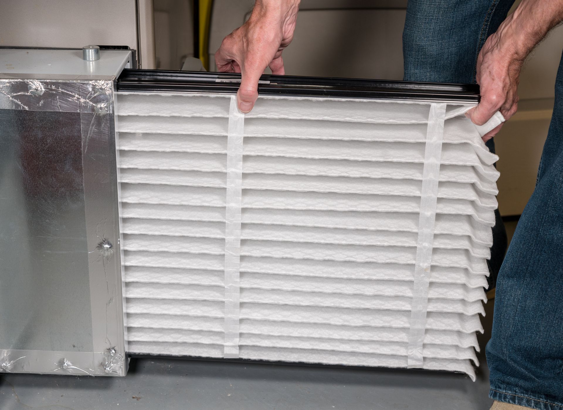 Person replacing an air filter in a furnace. Hands holding a new, pleated filter near the metal unit.