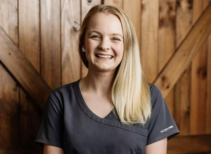 Woman with blonde hair wearing a grey shirt smiling in front of a wooden wall — Westlakes Veterinary Hospital in Fennell Bay, NSW