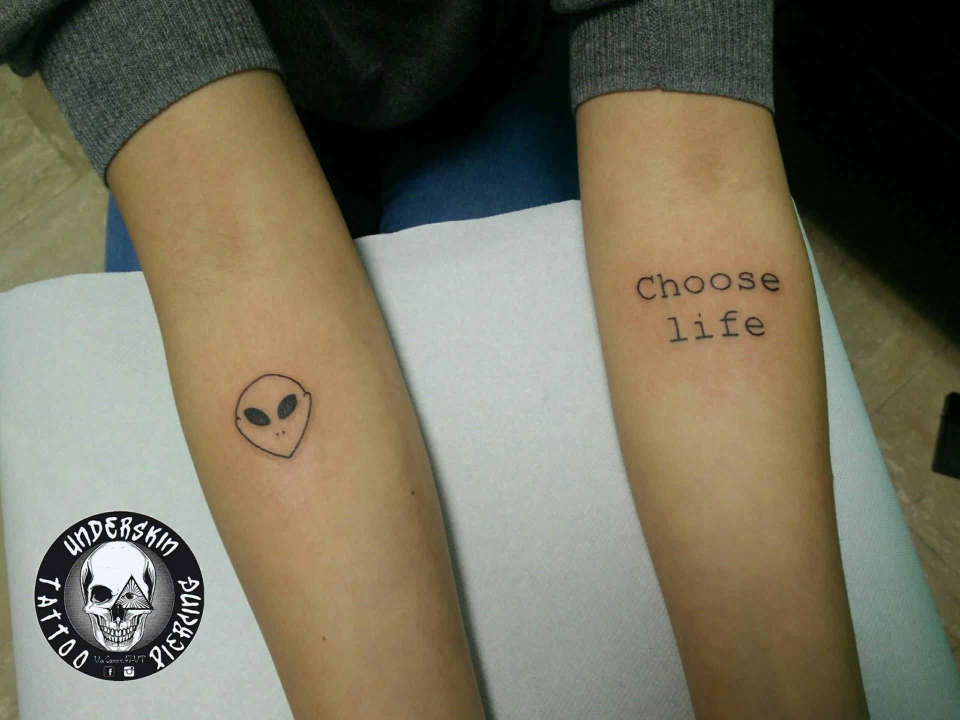 Tatuaggio di viso alieno e scritta sulle braccia