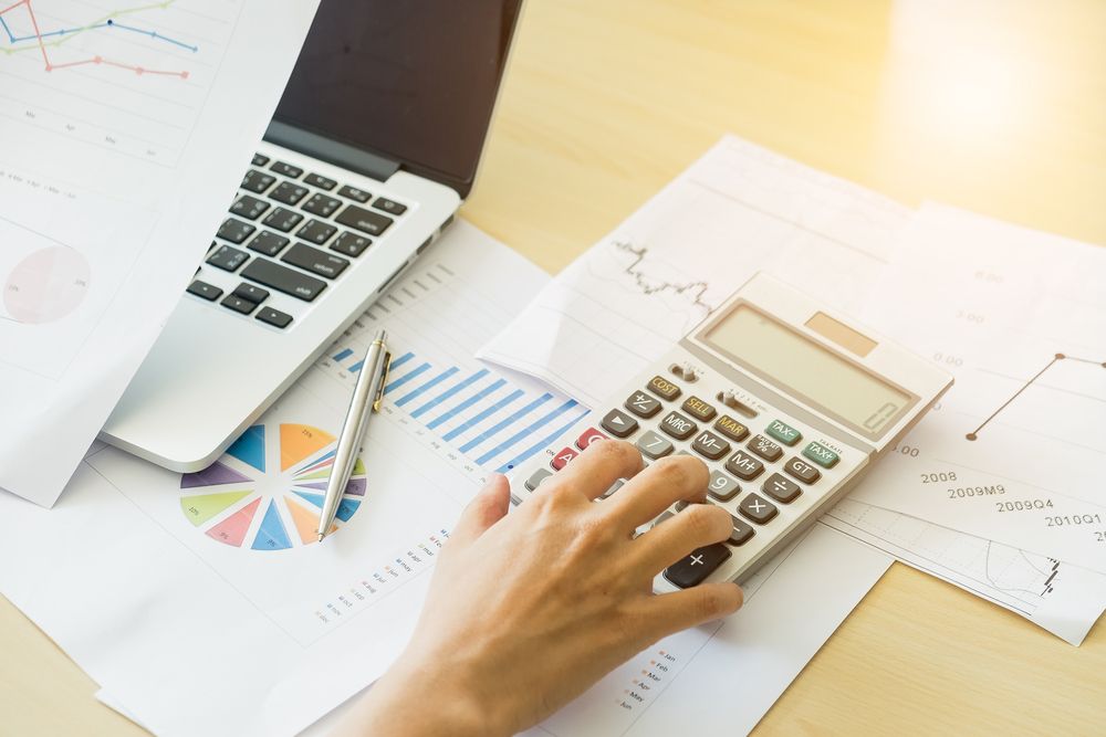 Person using a calculator at a desk with a laptop, graphs, and charts; calculating financial data. — Peterson Property Valuations In Caloundra West, QLD