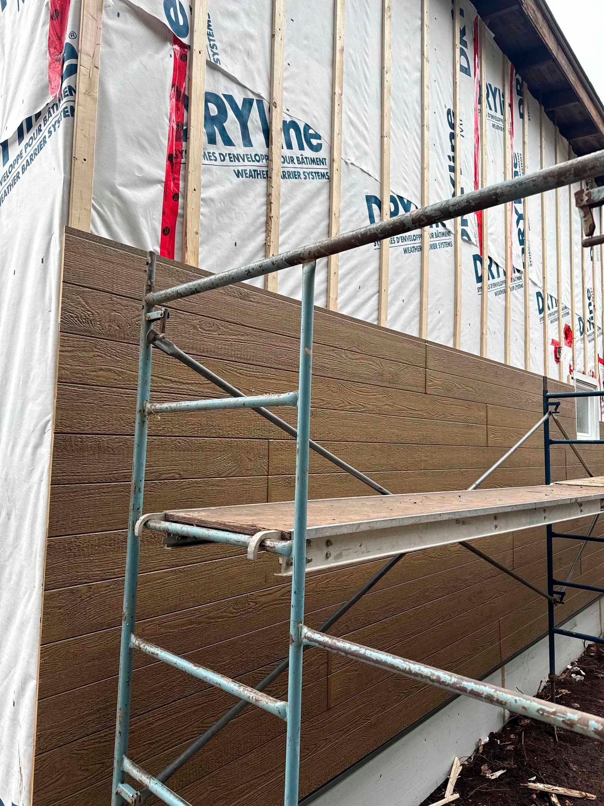 Chantier de construction avec bardage en bois et échafaudages adossés à un bâtiment, partiellement recouverts d'une bâche de protection blanche.