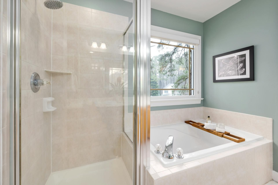 Modern bathroom with glass shower, white tub, and teal walls next to a window.