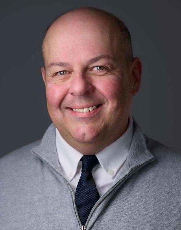 A bald man wearing a sweater and tie is smiling for the camera.