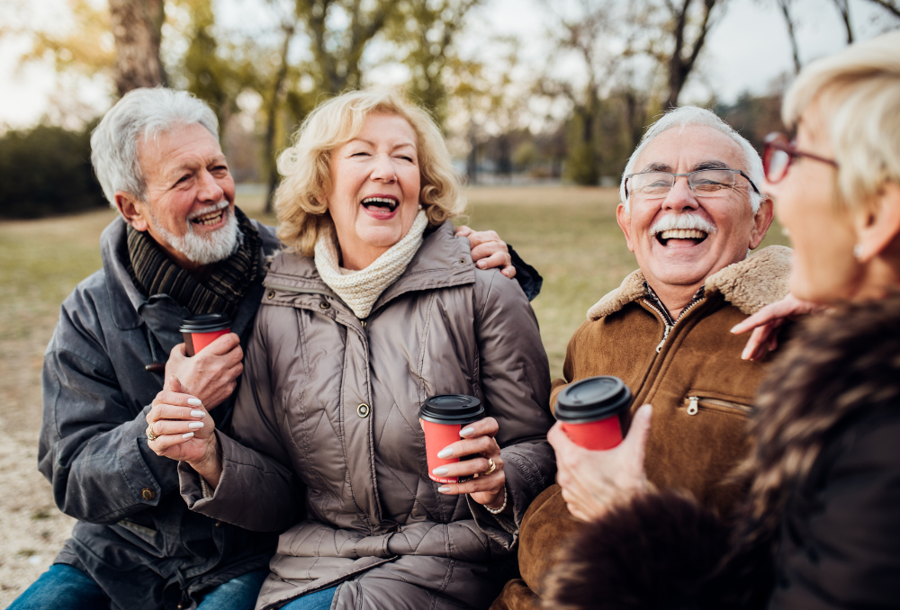 A group of elderly people are sitting on a bench in a park laughing and drinking coffee.
