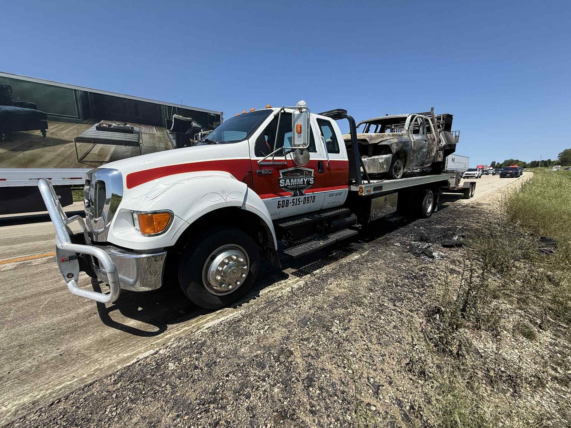 Tow truck carries a burnt vehicle on the side of a road under a blue sky.