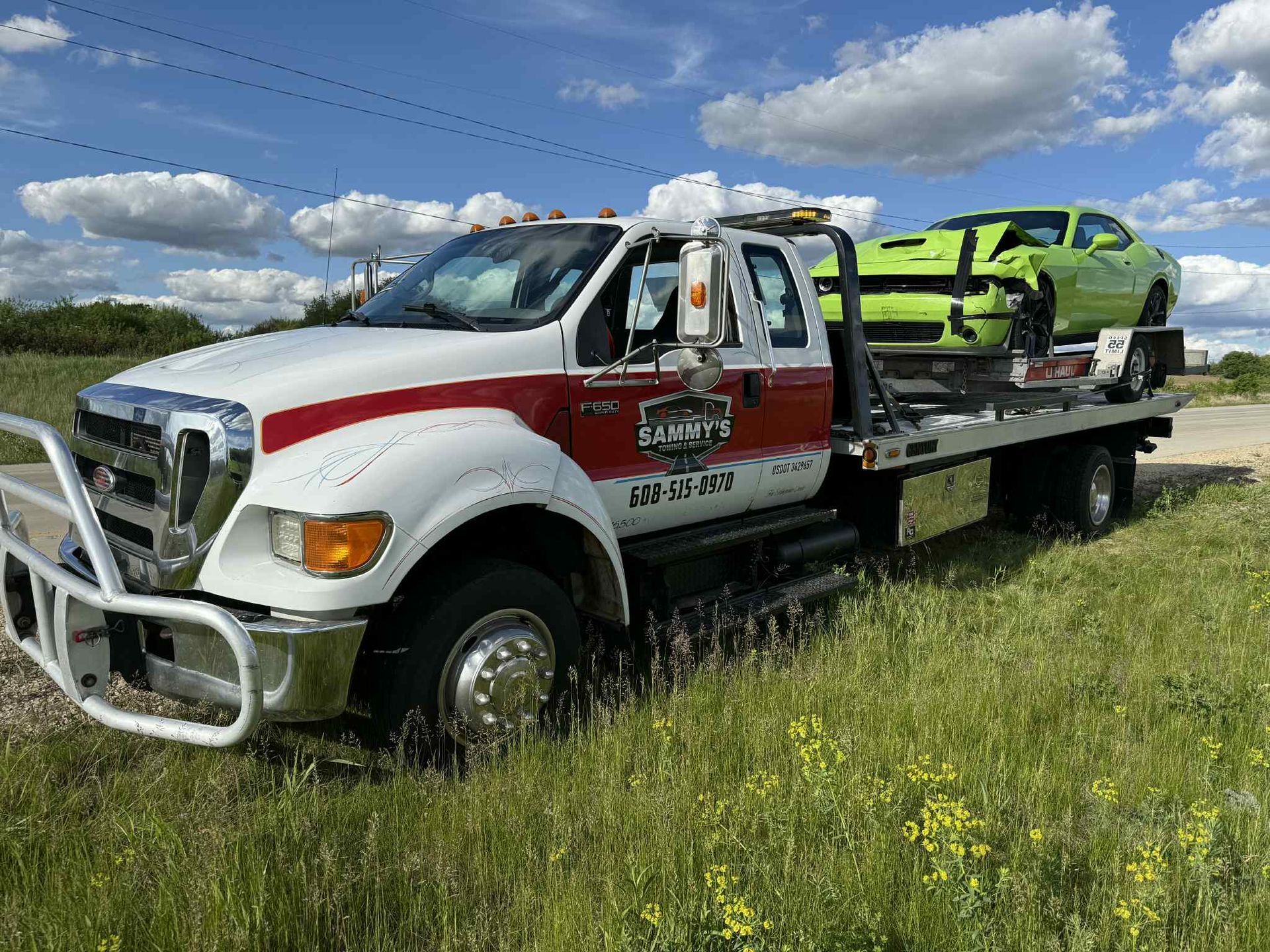 Tow truck with a green sports car on its flatbed, parked in a grassy area on a sunny day.