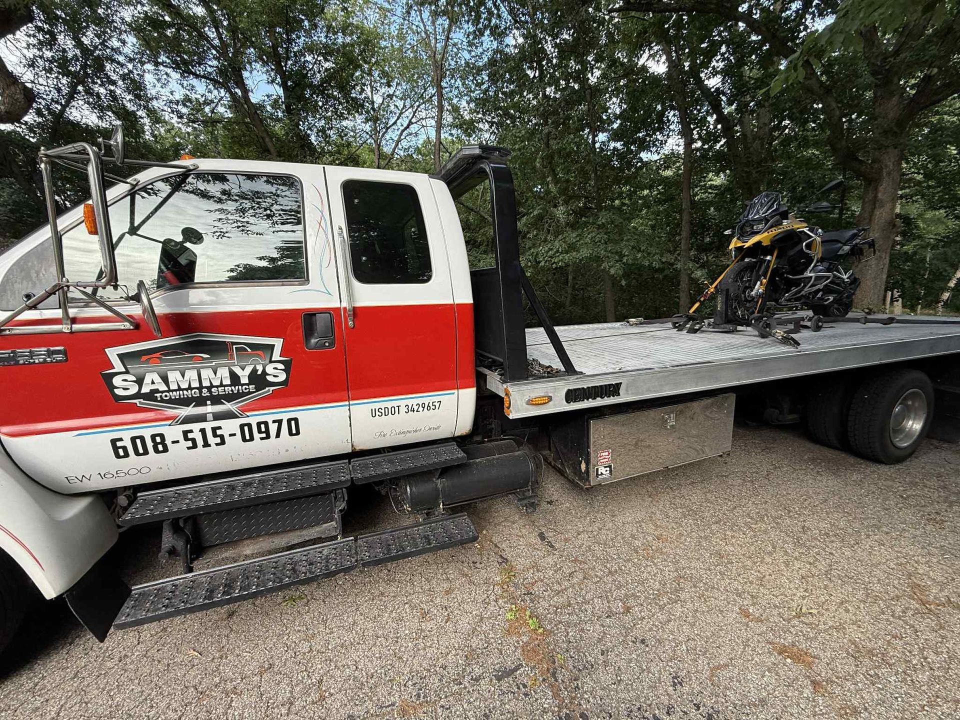 A white and red Sammy’s flatbed tow truck with a motorcycle secured on the back, parked on a gravel lot near trees.