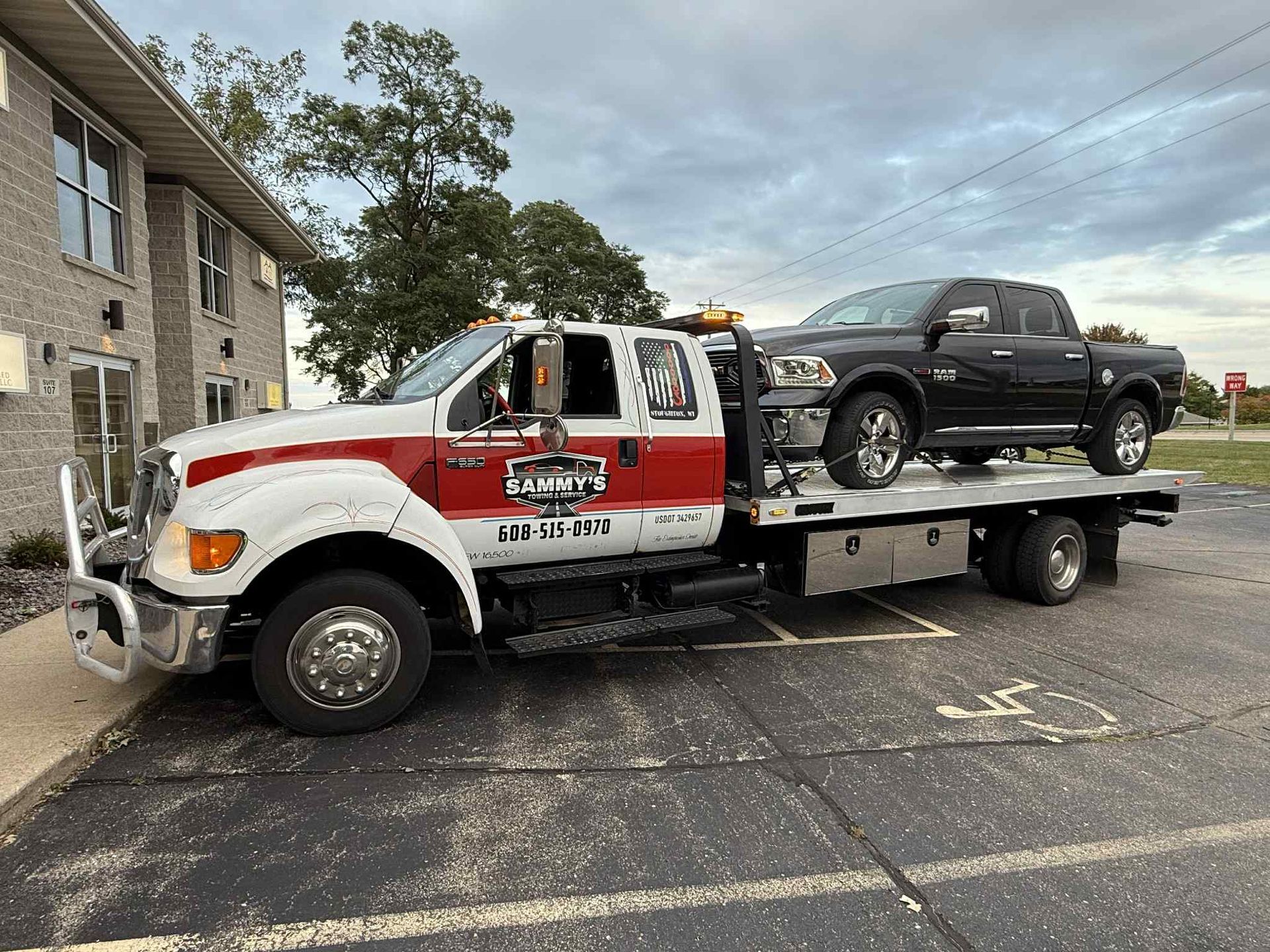 Tow truck with a black pickup truck loaded on the flatbed, parked in a lot next to a building.