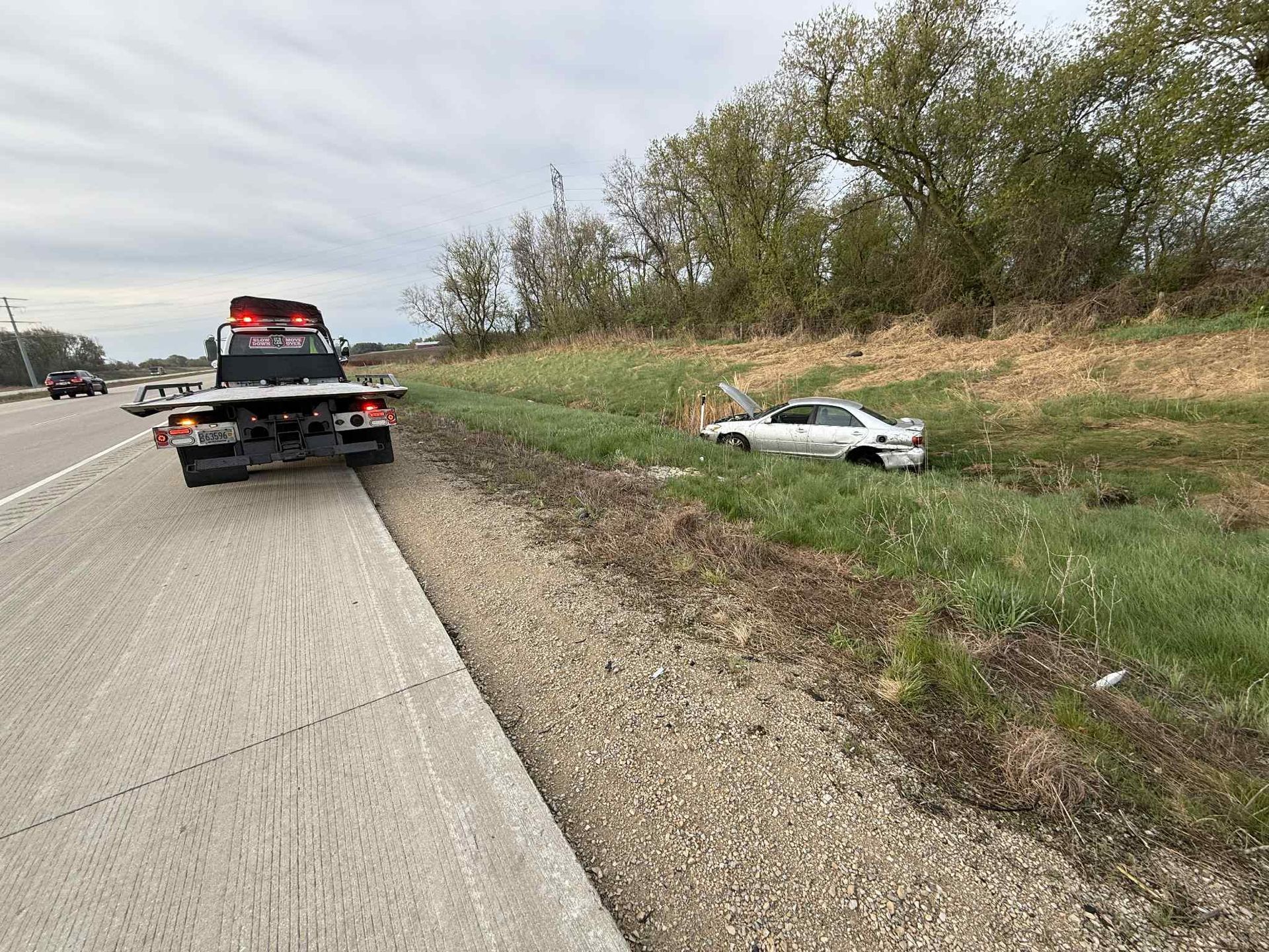 A silver car sits off the side of a highway in a grassy ditch, with a tow truck parked on the road nearby.