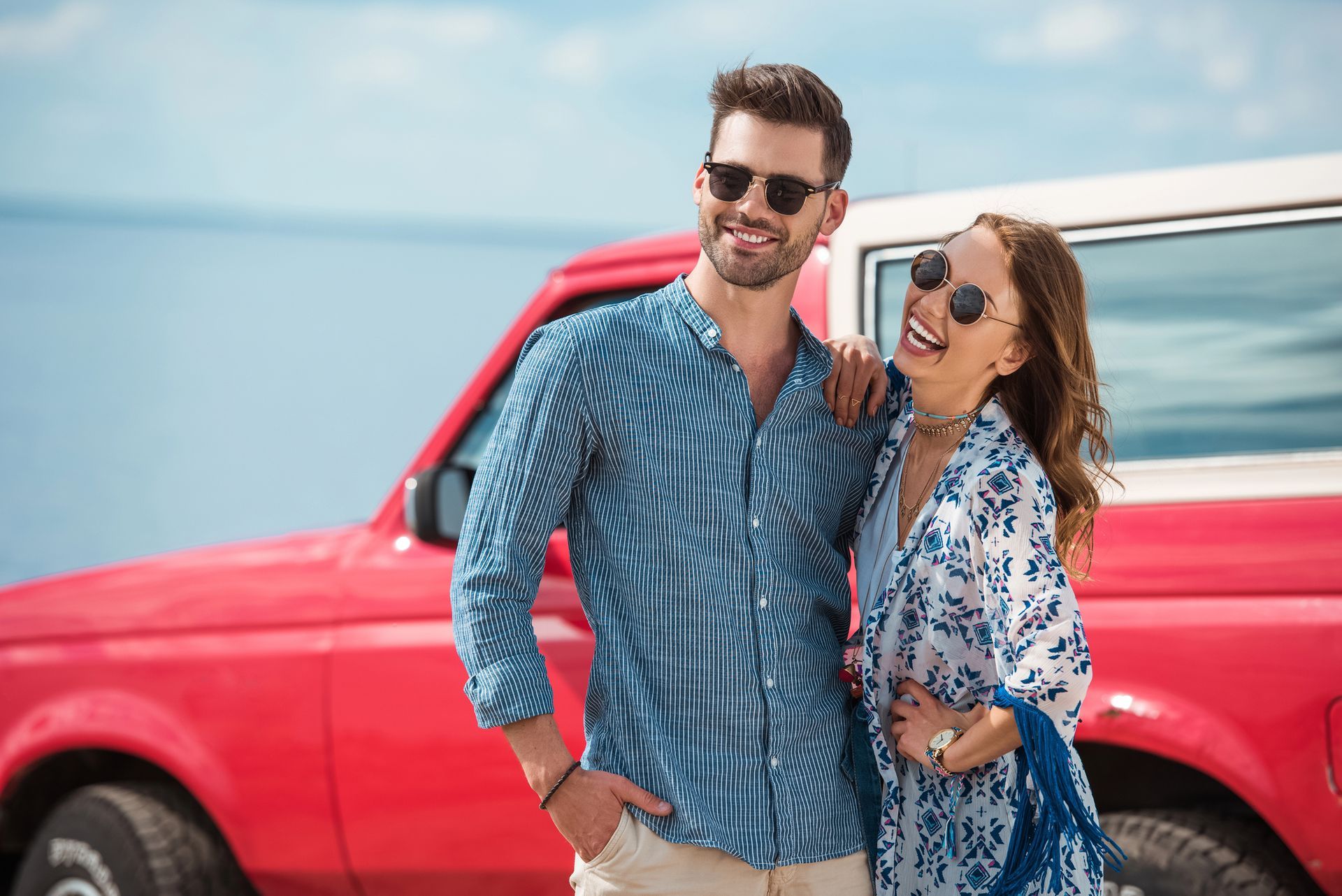 Una coppia con occhiali da sole sorride, vicino a un camion rosso vicino all'oceano.