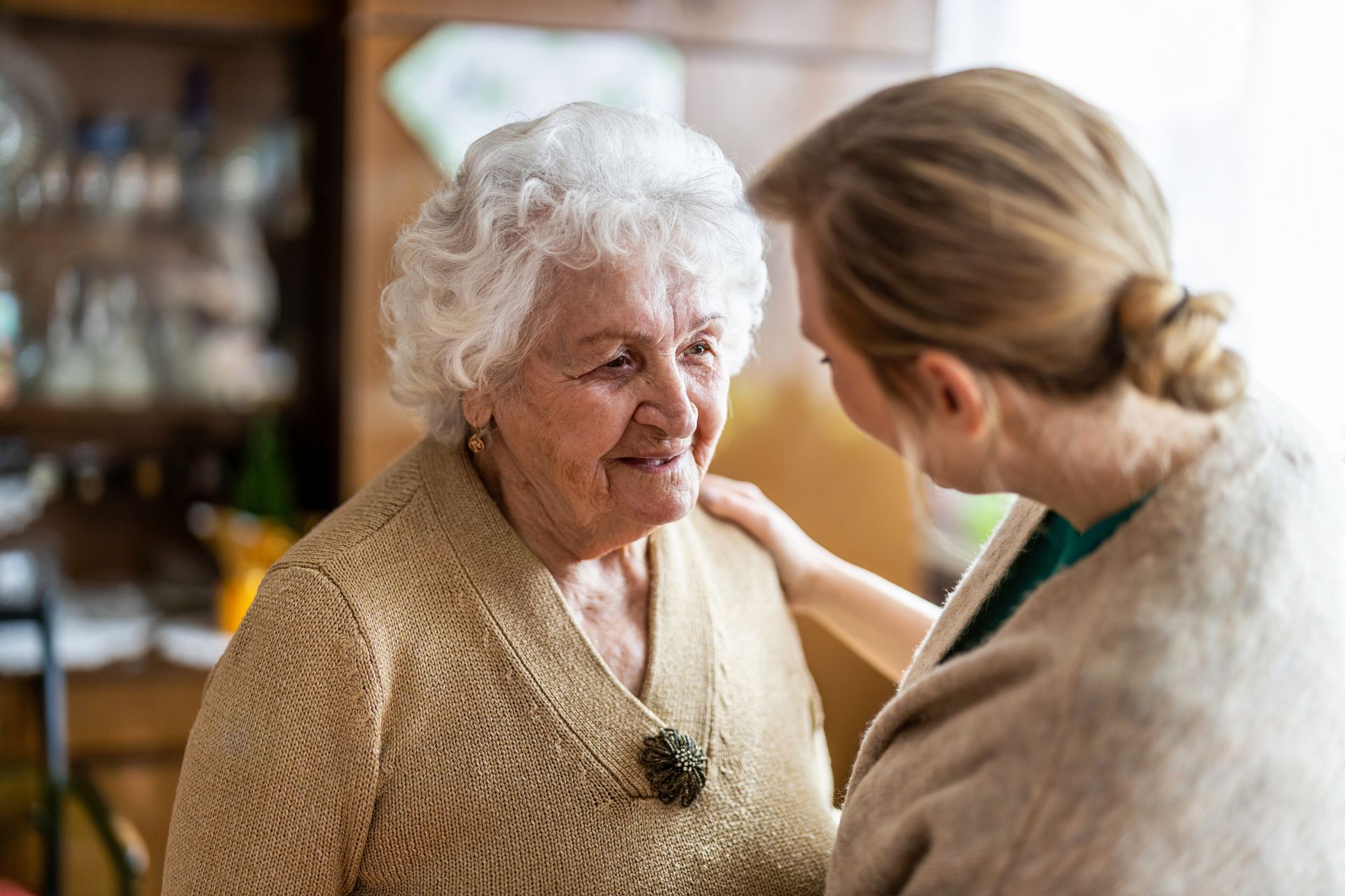 A woman standing close and touching an older woman’s shoulder inside a home interior.