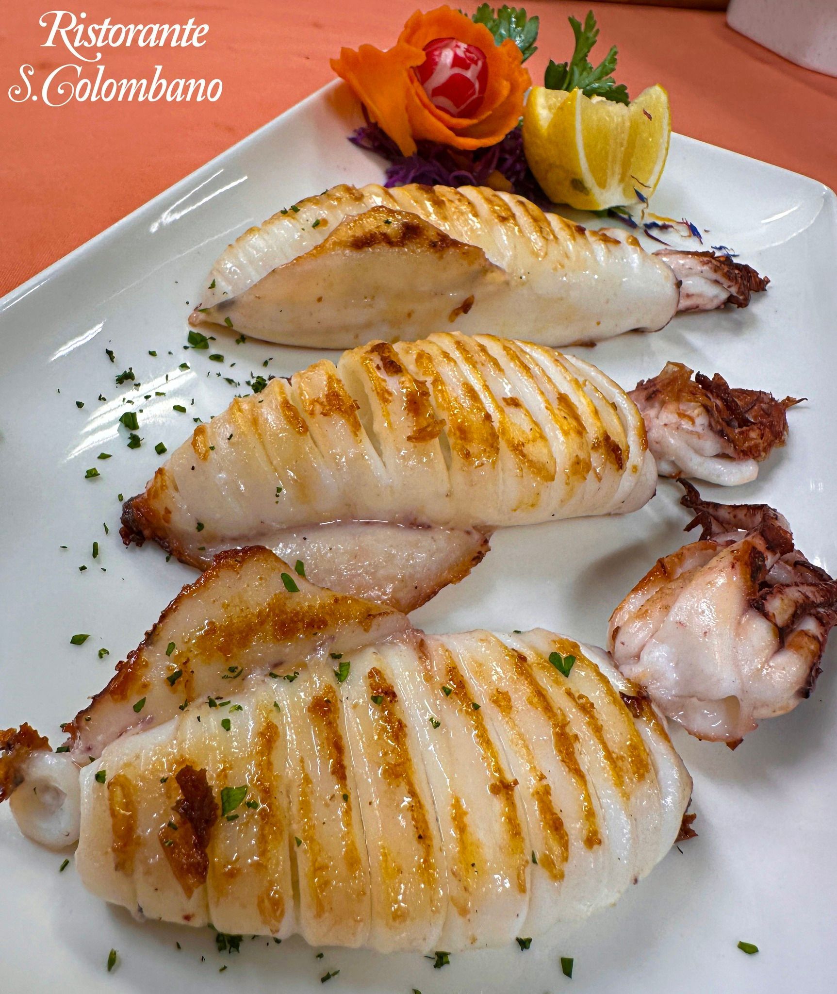 Un piatto con tre pezzi di calamari grigliati guarniti con prezzemolo, uno spicchio di limone e un intaglio decorativo di verdure.