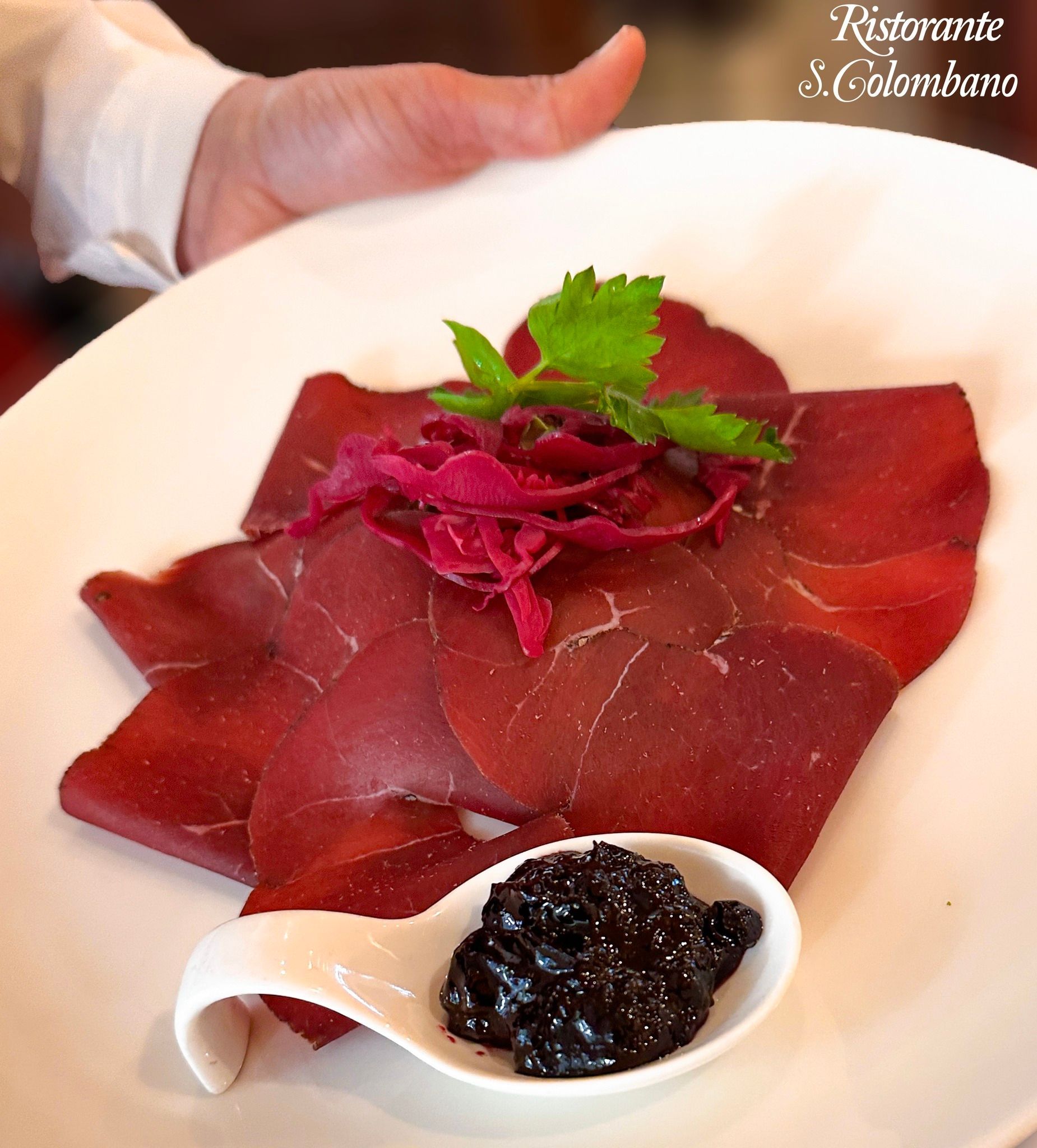 Una mano regge un piatto bianco di bresaola tagliata a fettine sottili e guarnita con verdure, servita con un cucchiaino di tapenade scura.