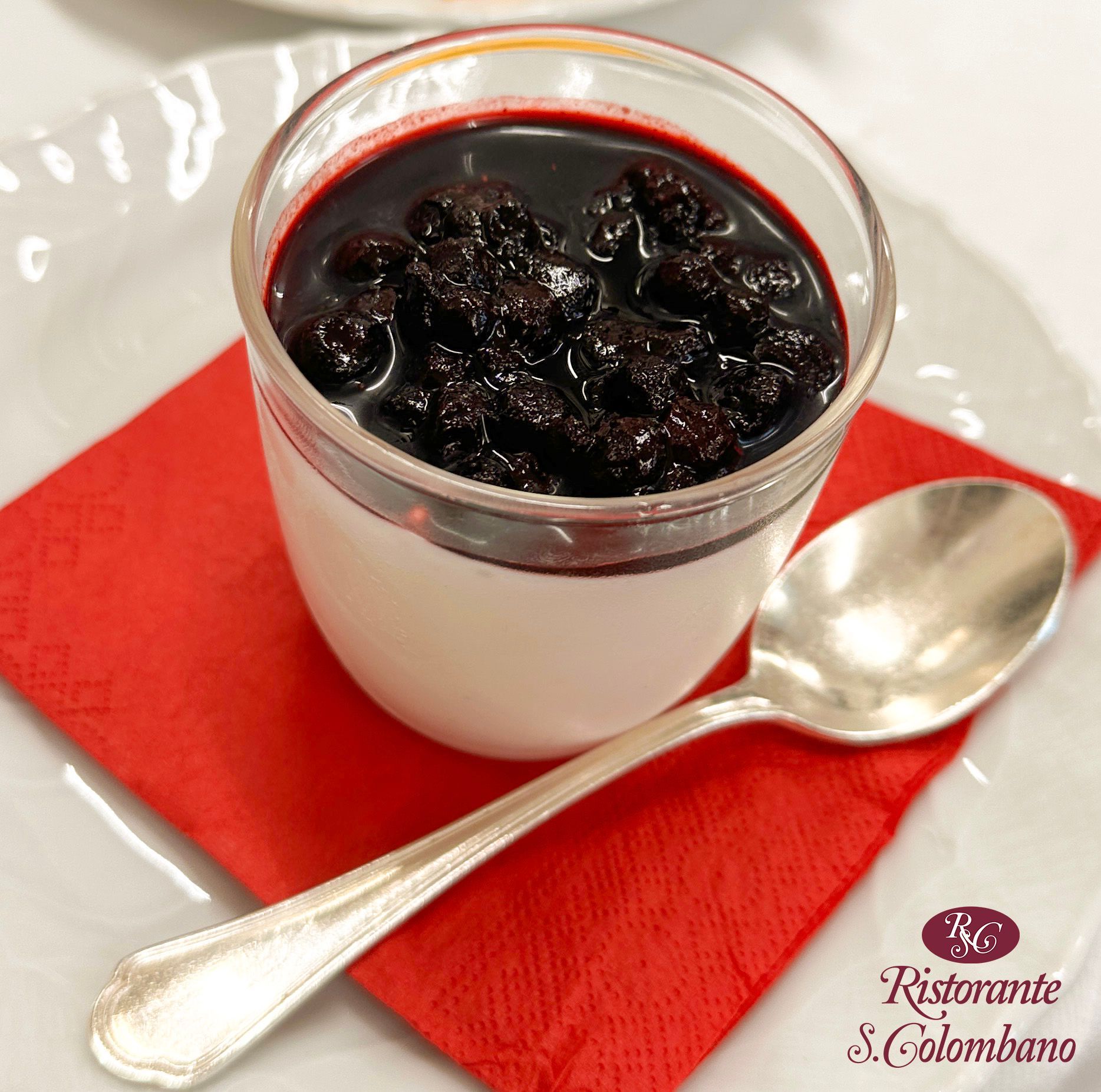 Una coppetta di panna cotta con composta di frutti di bosco, adagiata su un tovagliolo rosso con un cucchiaino d'argento su un piatto bianco.