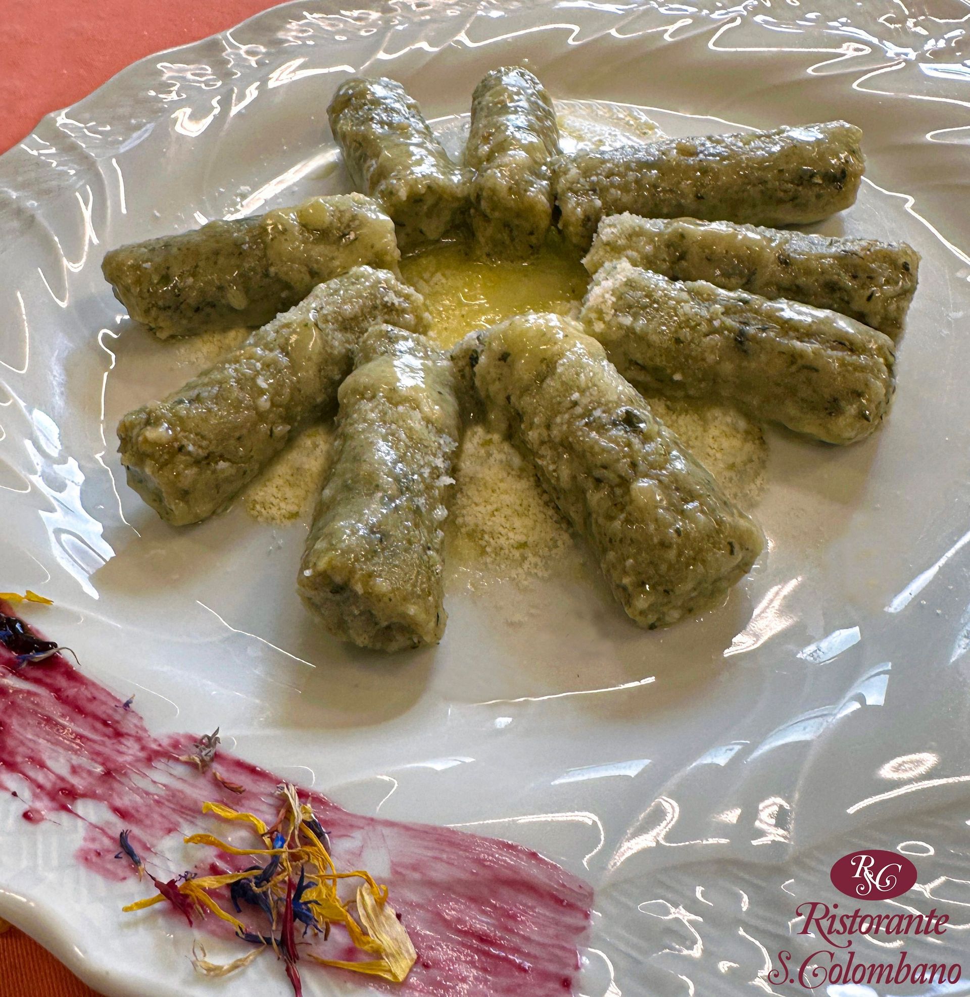 Gnocchi verdi cilindrici disposti in cerchio su un piatto bianco, con una spalmata di salsa ai frutti di bosco e fiori eduli a lato.