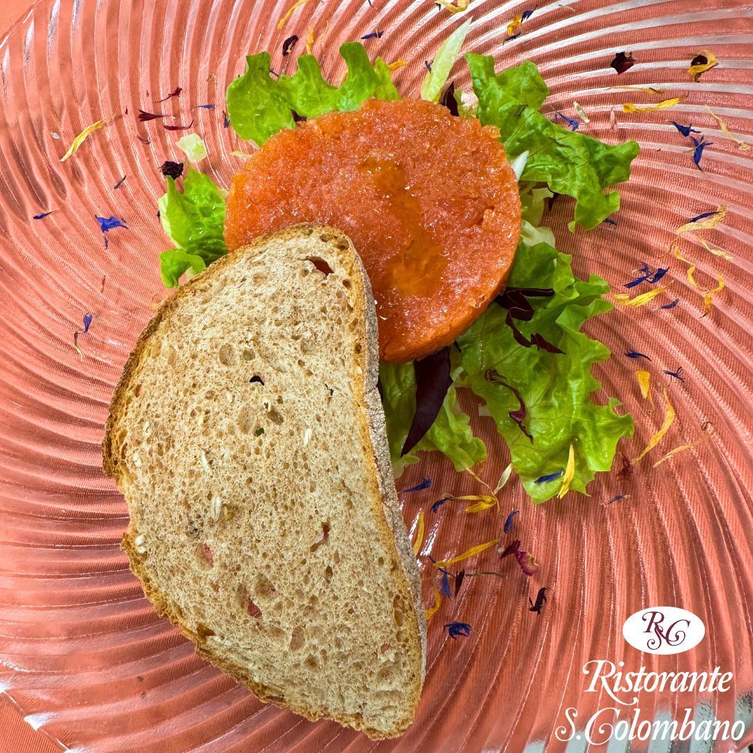 Una fetta di pane artigianale poggia accanto a un medaglione di tartare di salmone su un letto di verdure, servito su un piatto di vetro rosa a coste.
