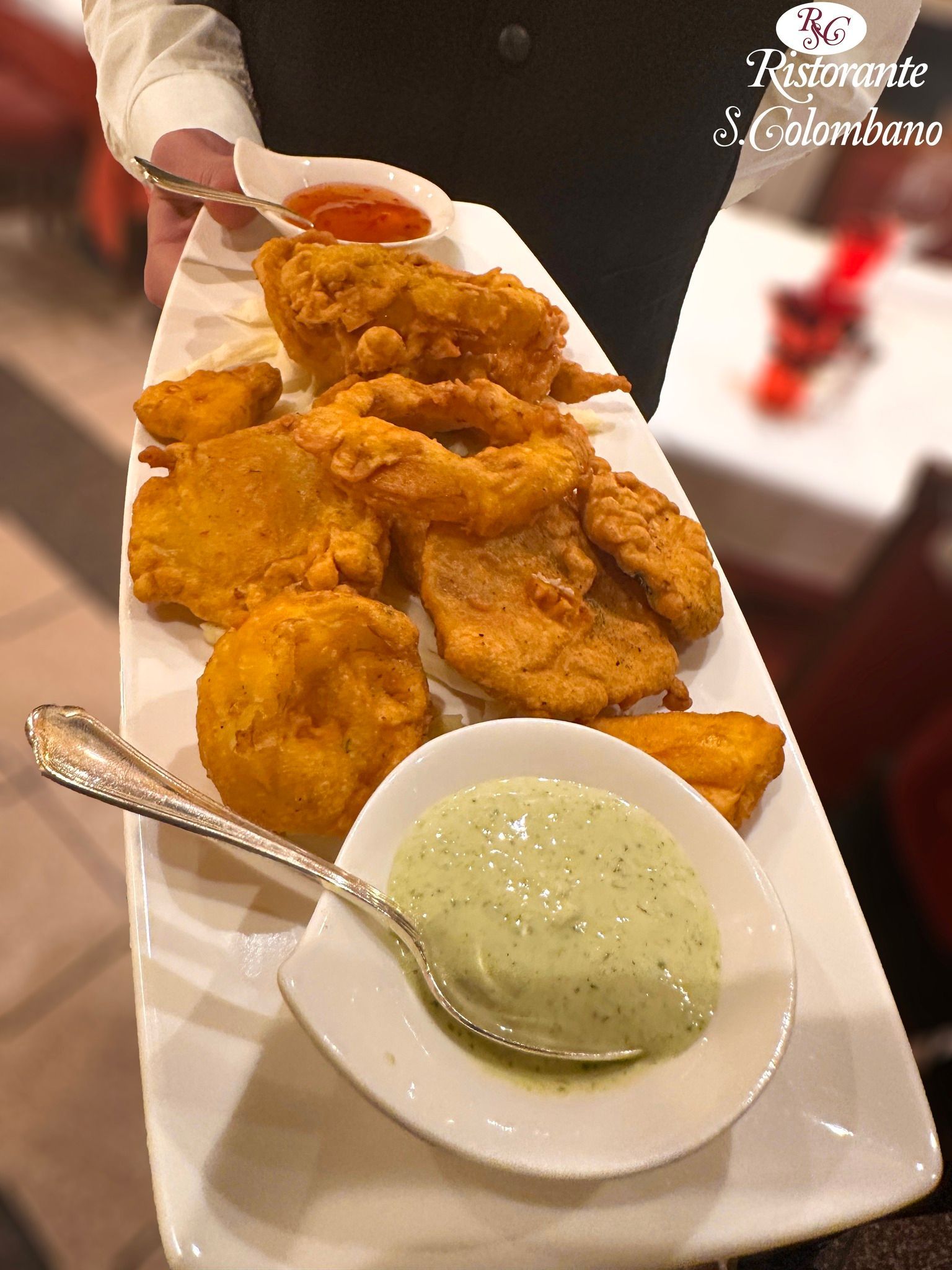 Un cameriere tiene in mano un vassoio rettangolare di antipasti fritti dorati con due piccole salse per intingere, una rossa e una verde.