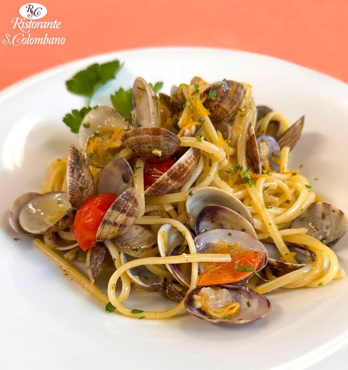Un piatto di linguine con vongole, pomodorini e una guarnizione di prezzemolo fresco su un letto di pasta arancione chiaro.