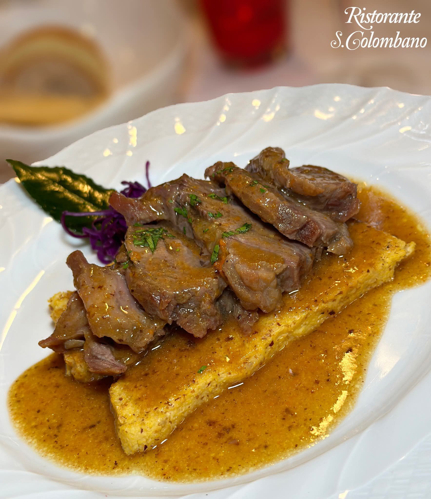 Fettine di tenera carne brasata servite su un rettangolo di polenta giallo dorata in una salsa saporita, su un piatto bianco.