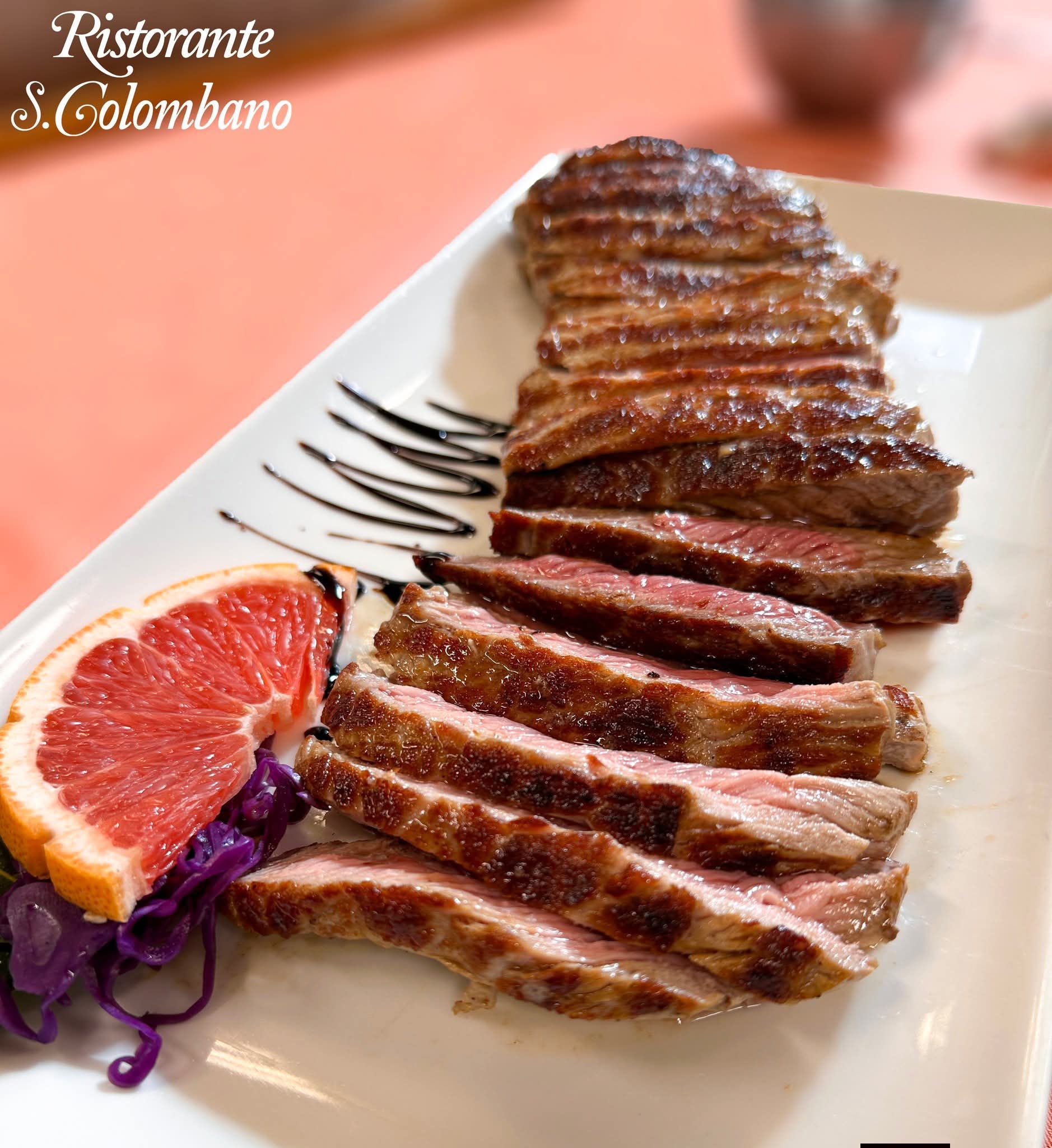 Bistecca grigliata a fette, servita su un piatto rettangolare bianco con una fetta di pompelmo e una guarnizione di cavolo rosso.