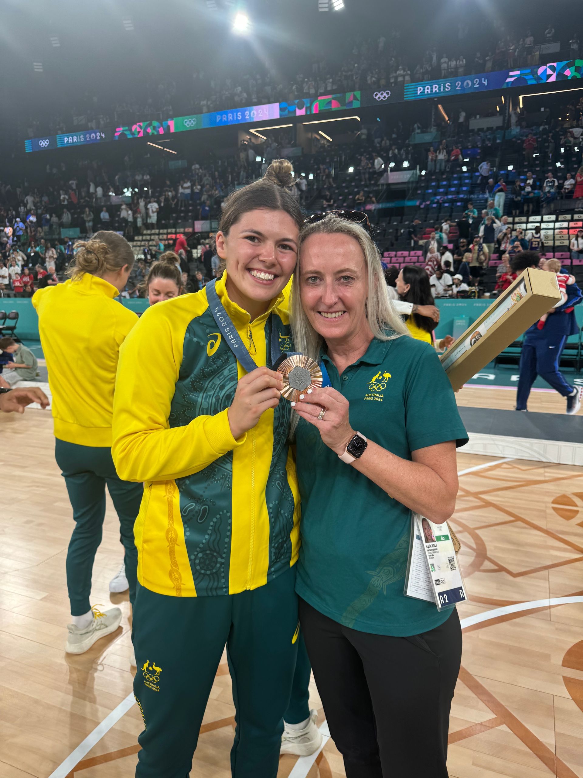 Two smiling people in Australian team uniforms holding a bronze medal on a sports court after an event.