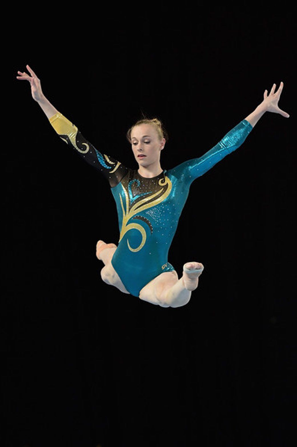 A female gymnast is jumping in the air with her arms outstretched.