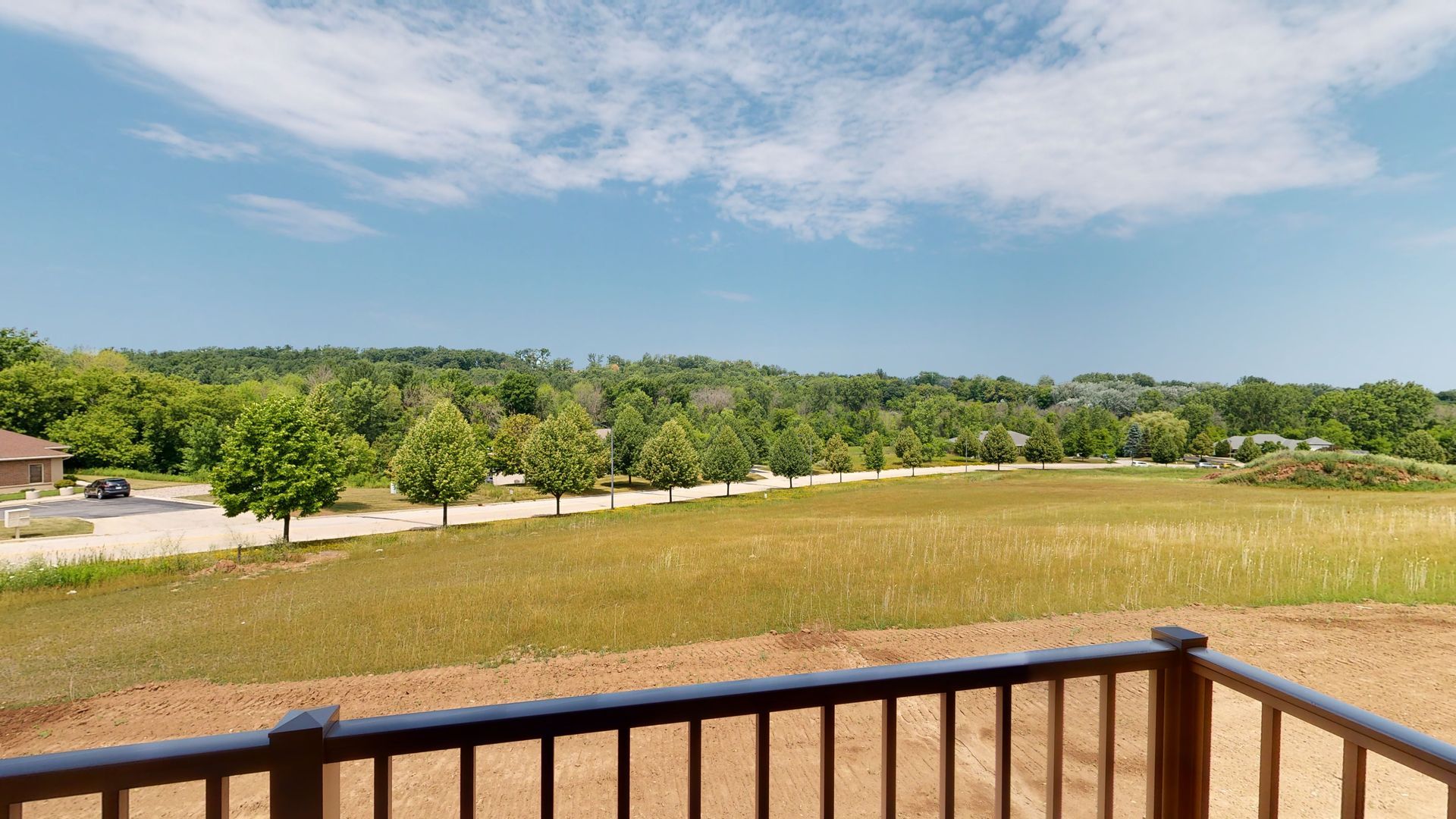 View from a wooden deck over a grassy field and tree line under a blue sky with scattered clouds