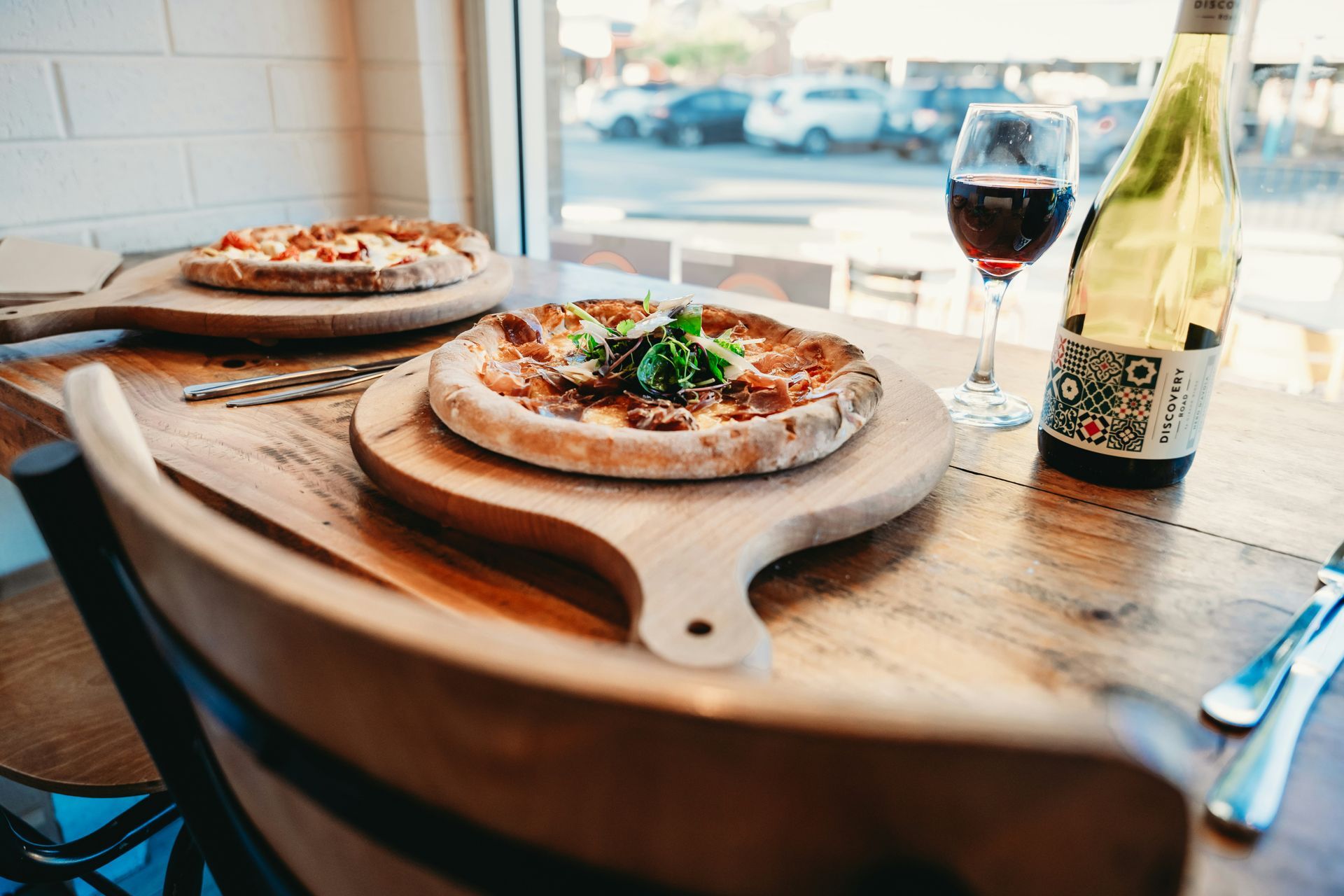 Two pizzas on wooden boards beside a wine glass and bottle by a windowed cafe table