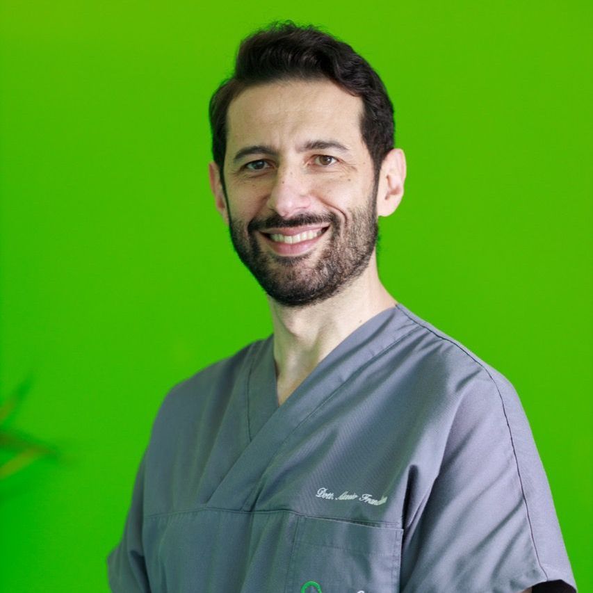 Un uomo con la barba indossa un camice e sorride davanti a uno sfondo verde.