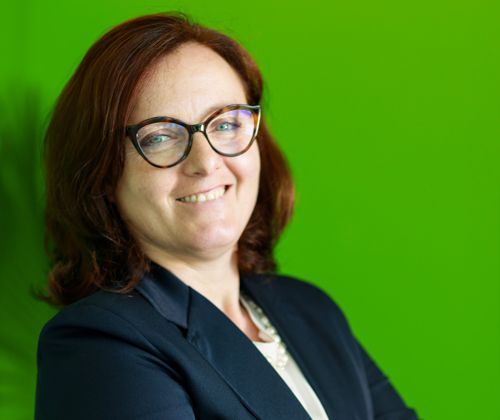 Una donna che indossa occhiali e un abito sorride davanti a uno sfondo verde.