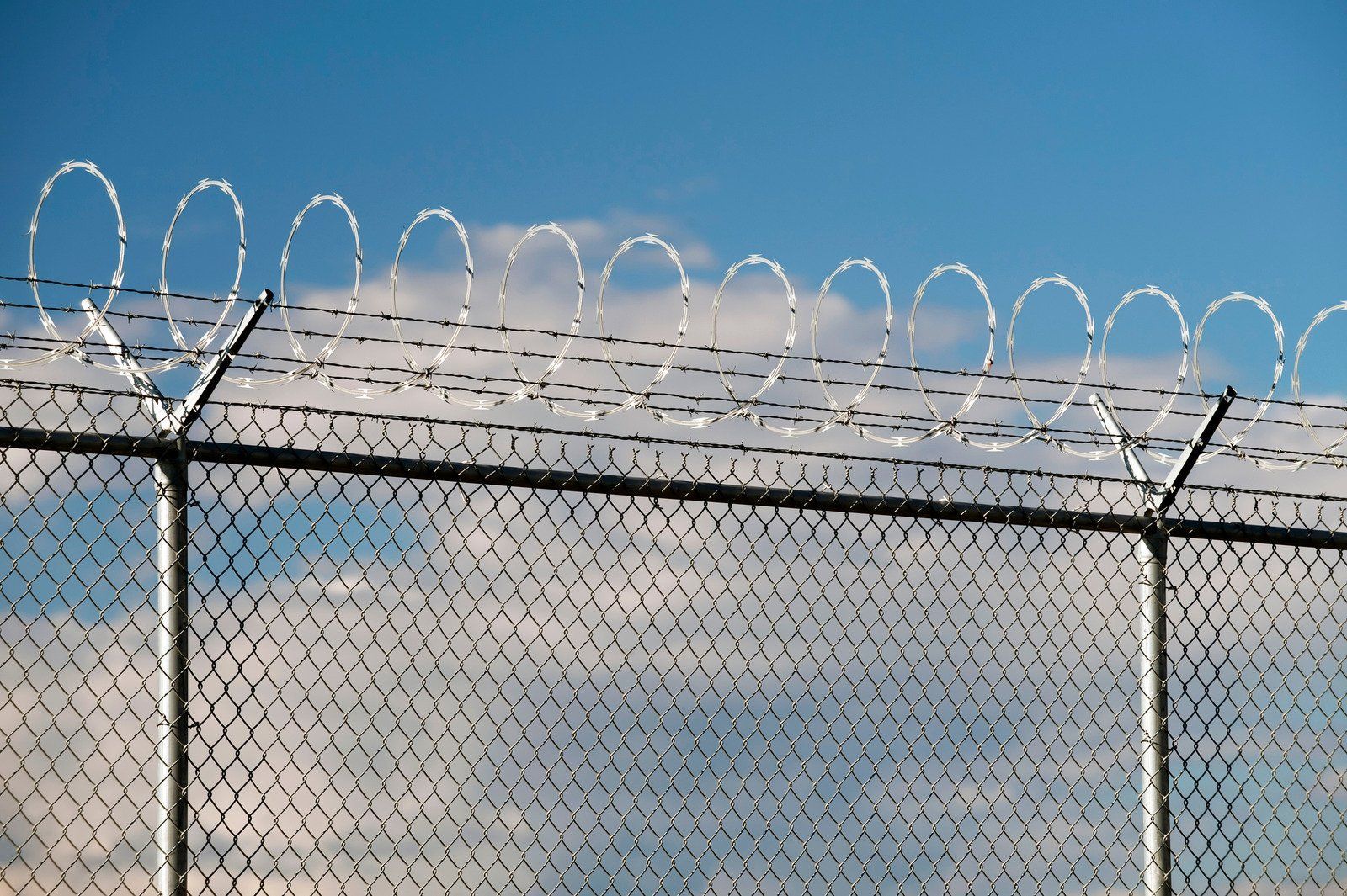 Chain-link fence topped with razor wire against a blue sky with clouds.