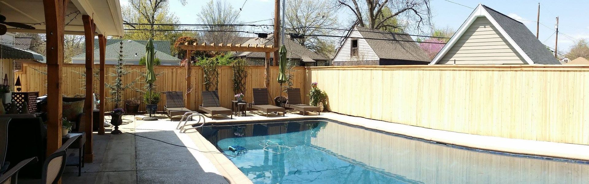 A backyard pool with lounge chairs under a wooden pergola, surrounded by a wooden fence, on a sunny day.